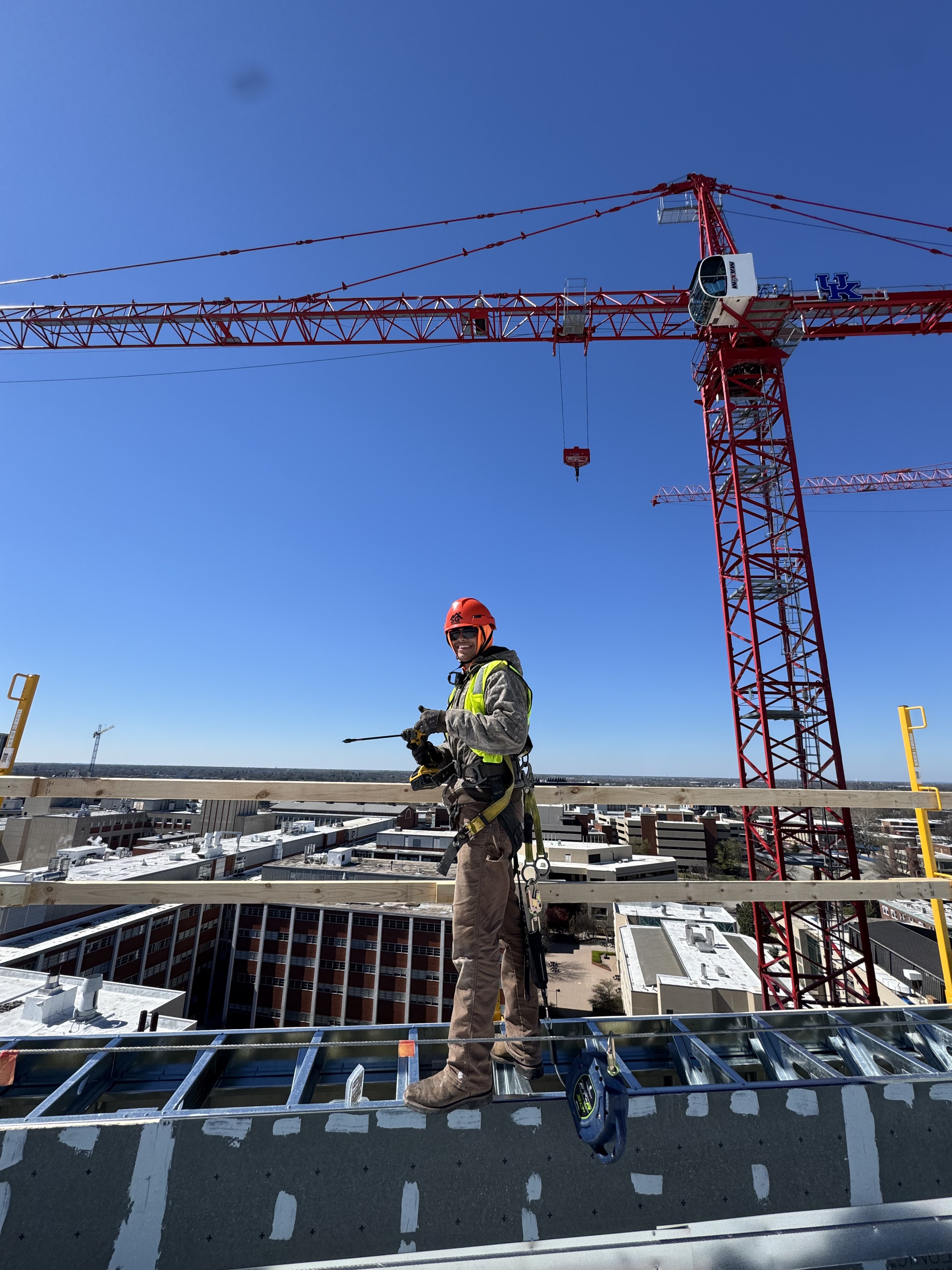 Worker wearing an orange hard hat, high-visibility vest, and safety harness on a construction roof beside a red crane.
